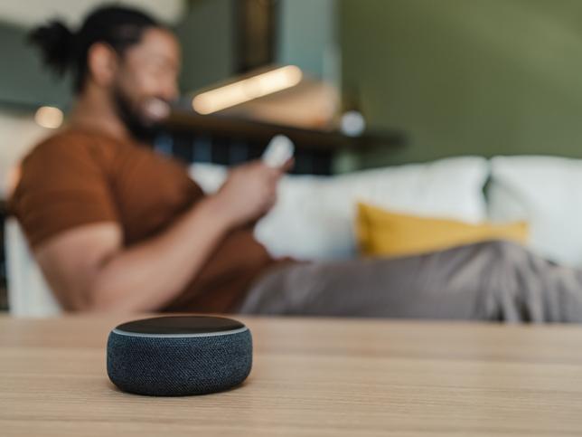 A smart speaker on a tabletop, and in an out-of-focus background a man holding a smartphone on a couch