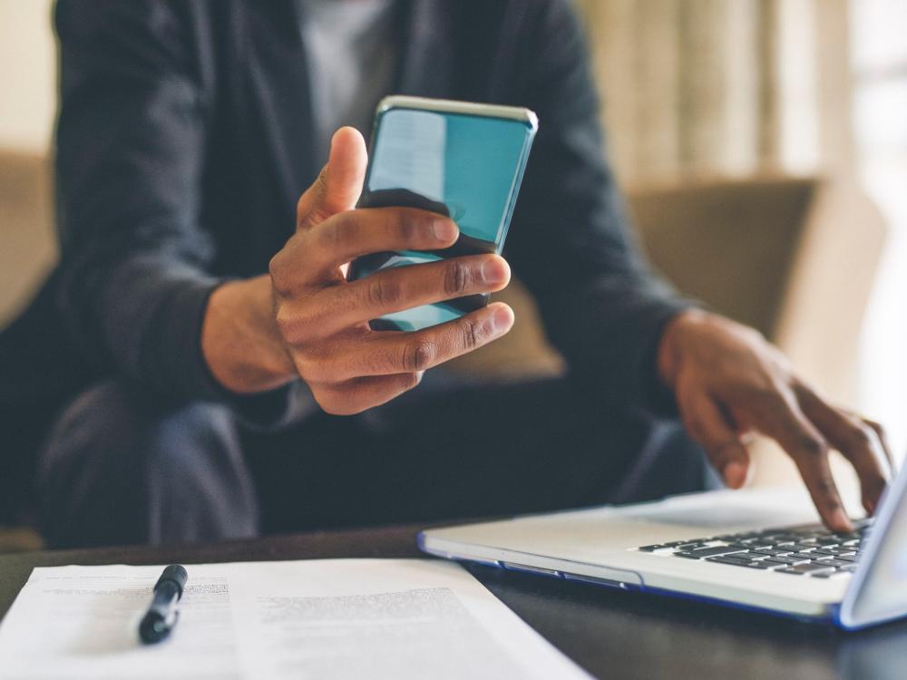 A man looks at his smartphone as he works on a laptop