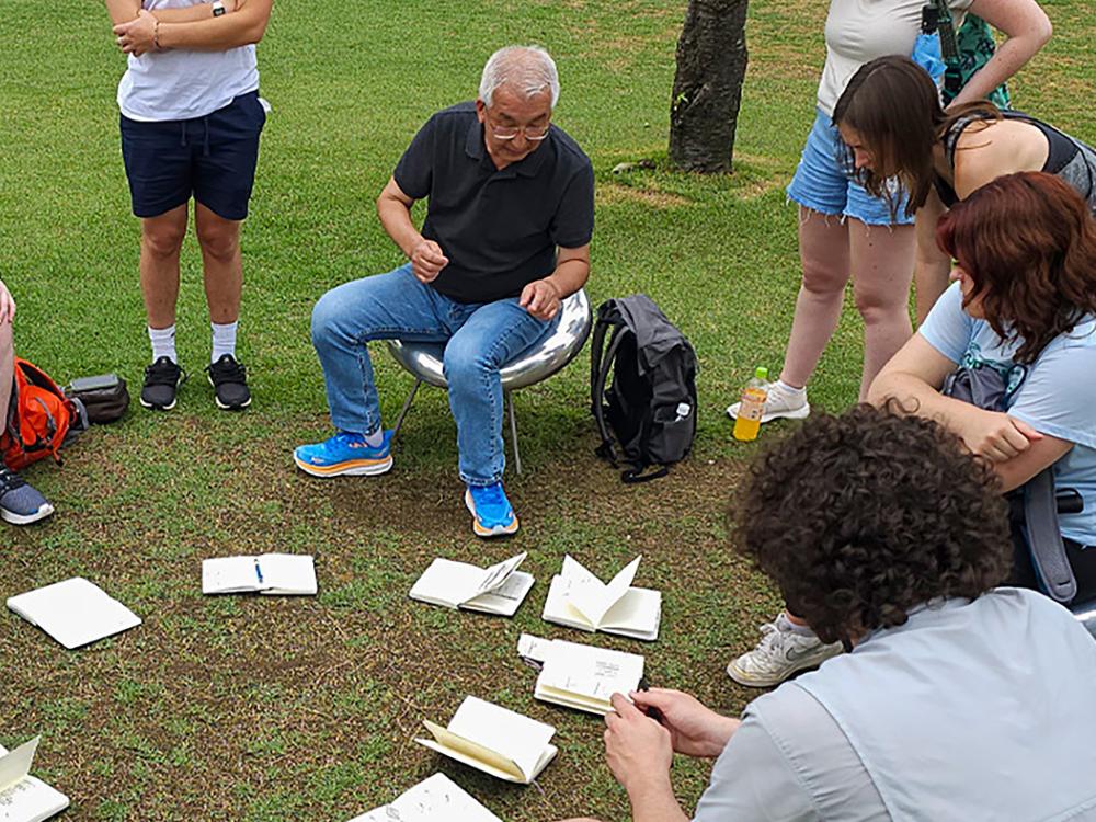 Katsuhiko Muramoto sitting among students outside with sketchbooks open on the ground in front of them.