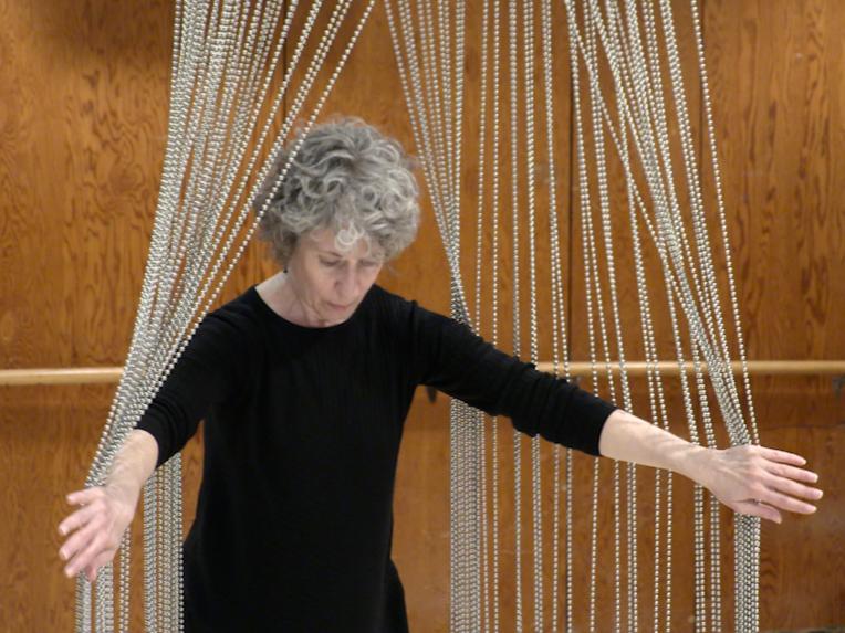 A woman with curly hair parts a beaded sculpture hanging from the ceiling.