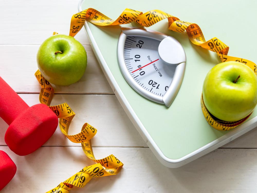 Green colored scale surrounded by two Granny Smith apples, yellow measuring tape next to small red-colored hand weights.