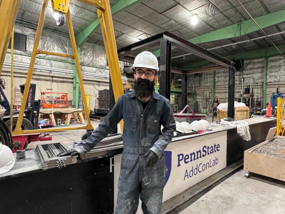 Ali Baghi stands in a hardhat and work jumpsuit in a workshop with a sign for the Penn State AddConLab behind him.
